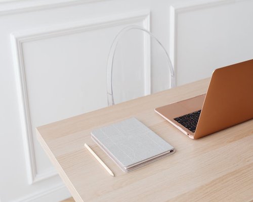 A laptop on a wooden desk next to a notebook in a bright room